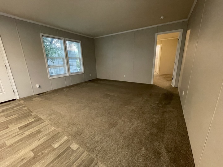 Empty room with beige carpet, light gray walls, and two windows with white blinds. Flooring transitions from wood-look vinyl to carpet. Door and hallway leading to other rooms are visible.