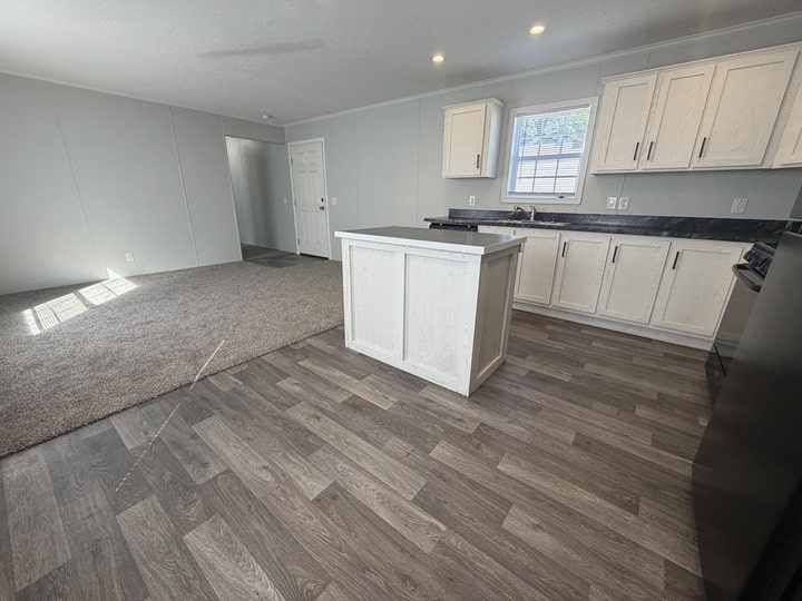 Open-plan kitchen and living area with light cabinets, a small island, dark countertops, and wood-look vinyl floors. The living space is carpeted, and a window lets in natural light. Walls are light gray.