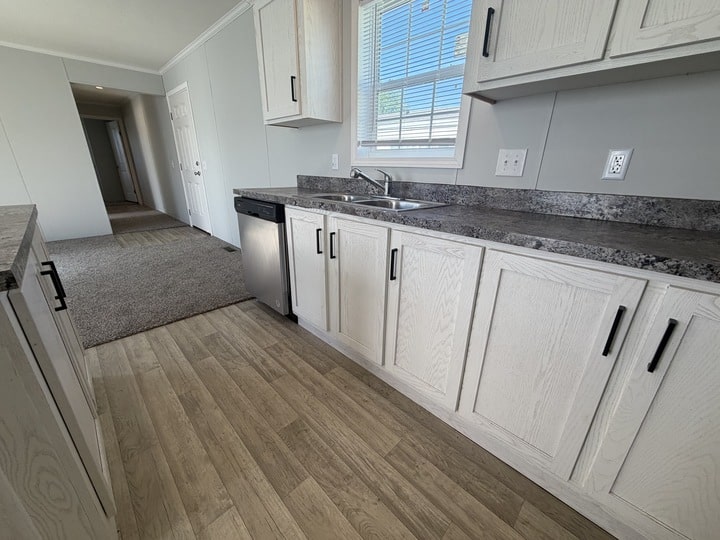 A modern kitchen with light wood cabinets, a stainless steel dishwasher, a double sink under a window, gray countertops, and light wood flooring adjoining a carpeted hallway.