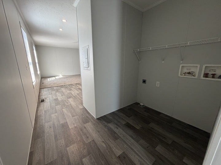 A hallway with gray wood flooring leads to a carpeted living area. On the right, there is a laundry nook with hookups and a wire shelf, all walls painted light gray. Sunlight enters through windows at the far end.