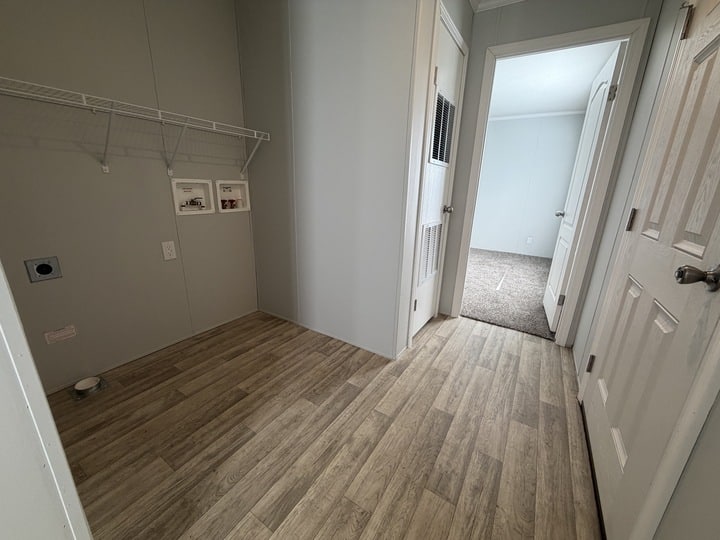 A small laundry room with wood-style flooring, empty wire shelf, and washer/dryer hookups; an open doorway leads to a carpeted room with white walls and a closed white door.