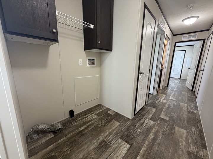A narrow hallway with wood-patterned flooring, white walls, dark wood cabinets, and several doors. An empty laundry nook with a vent pipe and wall shelf is visible on the left side.