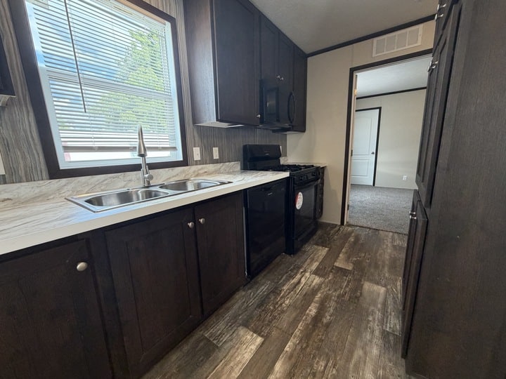 A modern kitchen with dark wood cabinets, a double sink under a window with blinds, a black stove and microwave, and wood-look flooring. An open doorway leads to a carpeted room.