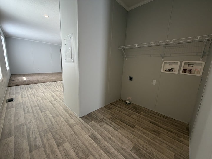 A laundry area with wood-look flooring, a white wire shelf, and hookups for a washer and dryer. The space opens into a carpeted living area with large windows along the left wall.