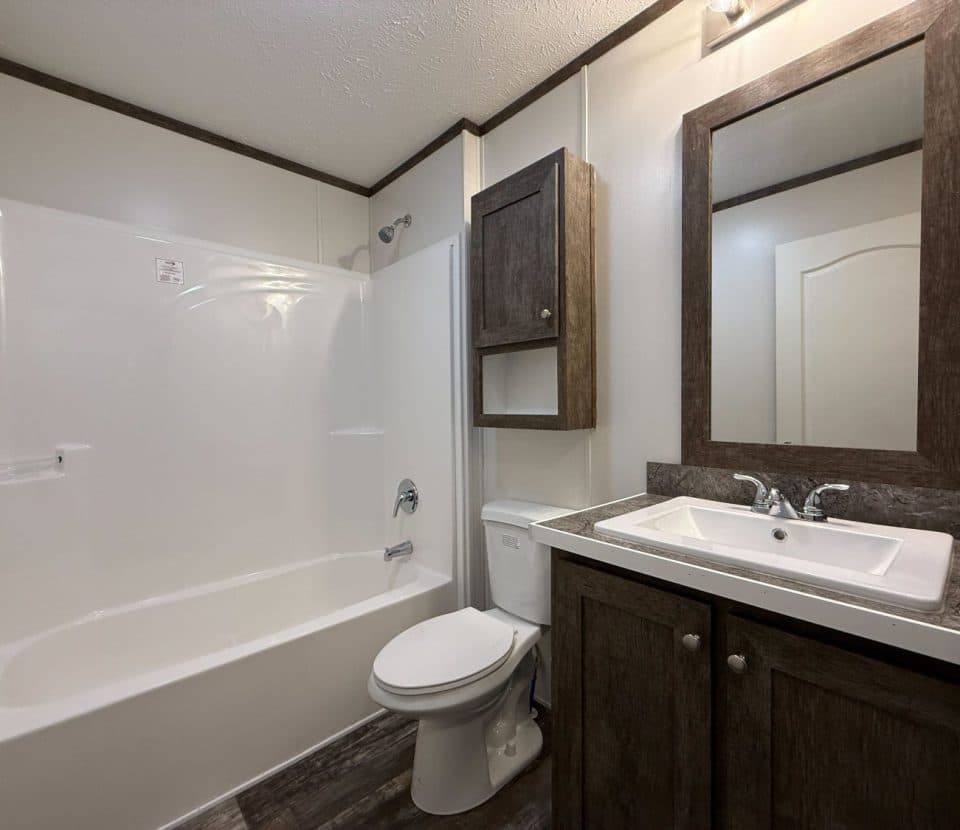 A modern bathroom with a white bathtub and shower, a toilet, and a dark wood vanity with a sink, matching cabinet, and a large mirror above the sink. The floor has a wood-like finish.