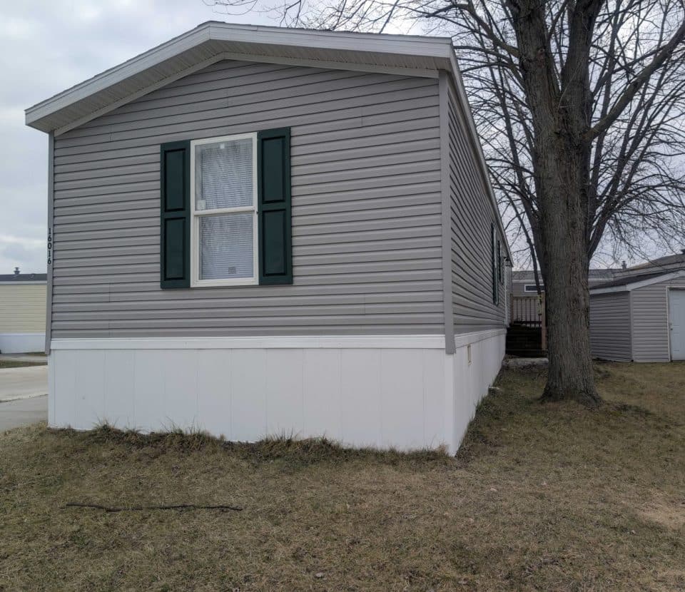 A gray mobile home with green shutters and white trim sits on a patch of dry grass near a leafless tree, with another mobile home and a small shed visible in the background.