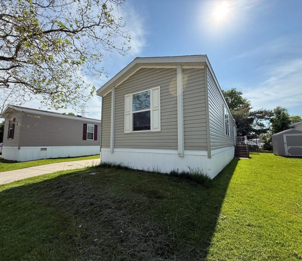 A beige mobile home with white trim and shutters sits on a grassy lawn under a sunny blue sky. A tree and another mobile home are visible nearby. Shadows are cast on the grass.