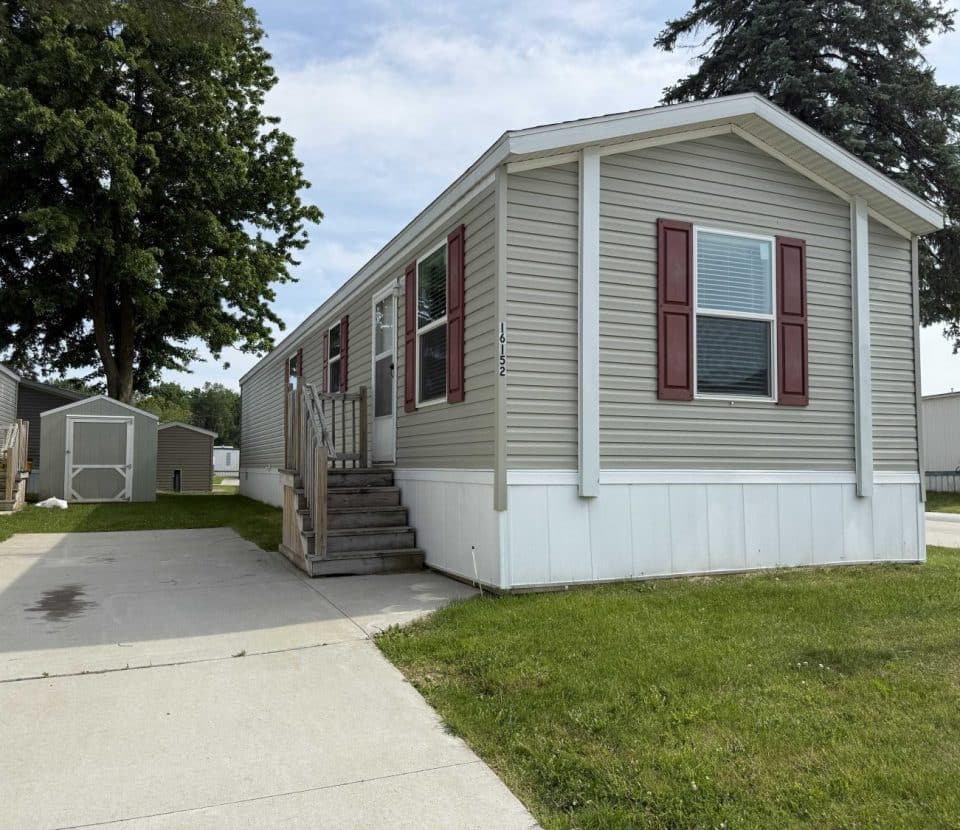 A beige mobile home with red shutters and a small wooden porch sits on a lot with a concrete driveway, green grass, and trees in the background. A white shed is visible behind the home.
