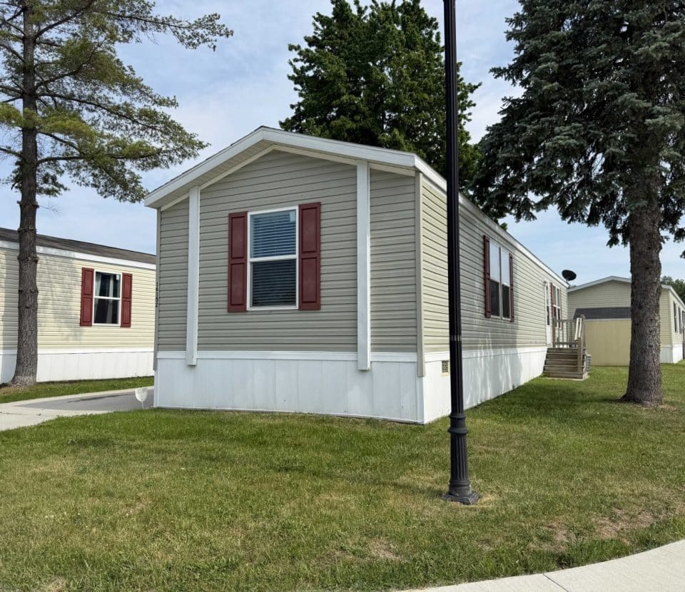 A beige mobile home with red shutters sits on a small grassy lot, bordered by a sidewalk and a street lamp. Other similar homes and trees are visible in the background under a blue sky.