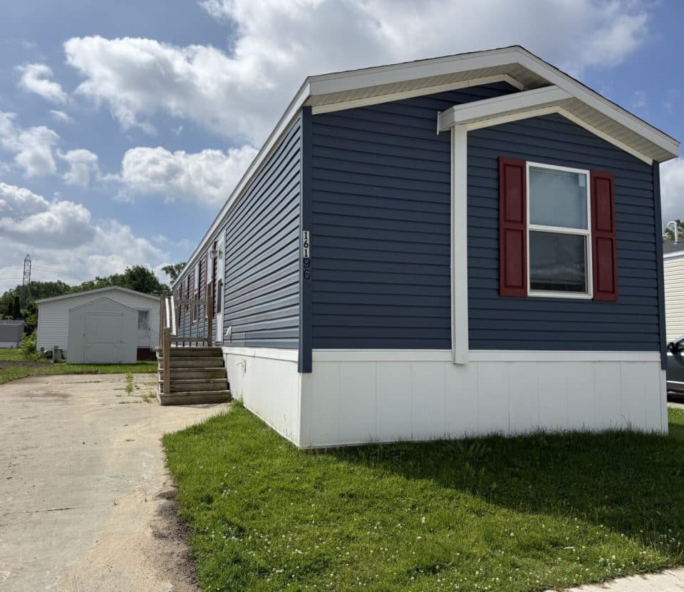 A blue manufactured home with red shutters and white trim sits on a concrete driveway with green grass on the side, under a partly cloudy sky. A small shed and parked cars are visible in the background.