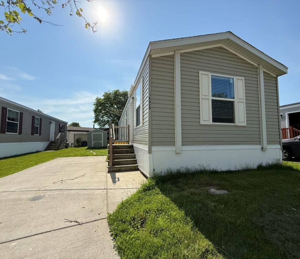 A small beige mobile home with white trim sits on a grassy lot, featuring a short stairway to the entrance and a concrete driveway. Other similar homes and a car are visible nearby under a clear blue sky.