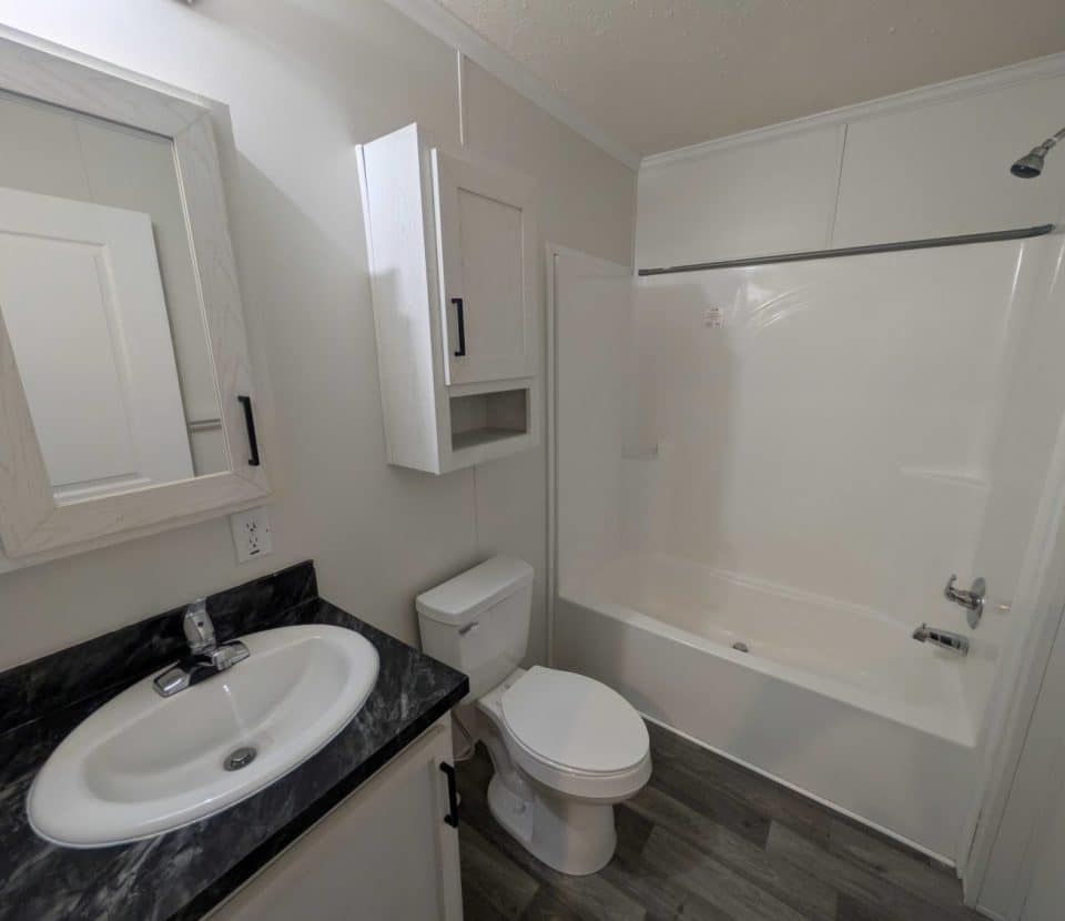 A small, modern bathroom with a sink and vanity, toilet, and a bathtub-shower combo. The walls are light-colored, the floor is gray wood-style, and there are white cabinets above and next to the sink.