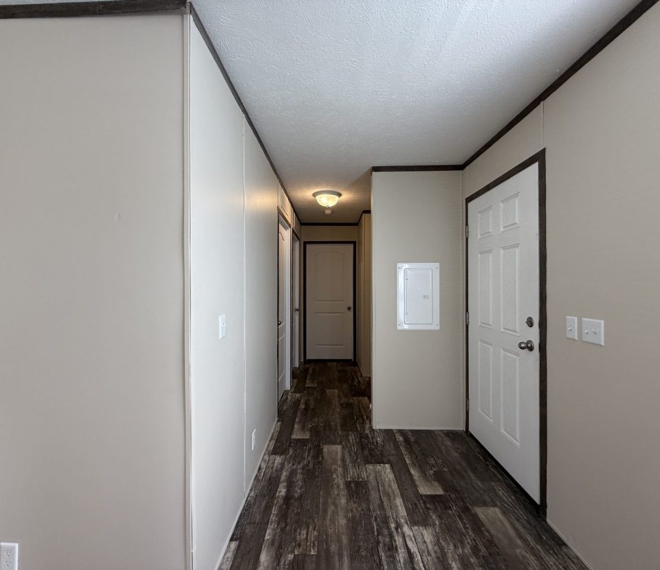 A hallway with beige walls, dark trim, wood-patterned flooring, a white door on the right, and a ceiling light; another closed door is visible at the end of the hall.