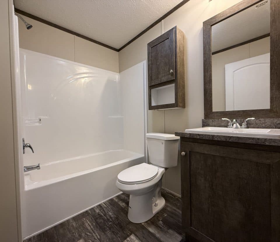 A modern bathroom with a white bathtub-shower combo, toilet, and a dark wood vanity with a matching mirror and cabinet, all set on a wood-look floor.