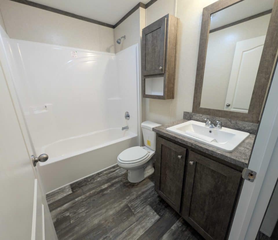 Modern bathroom with a bathtub-shower combo, toilet, and sink vanity with a mirror. Cabinets above and below the sink are wooden. The floor has a dark wood-like finish. Walls are light-colored. Door partially open.