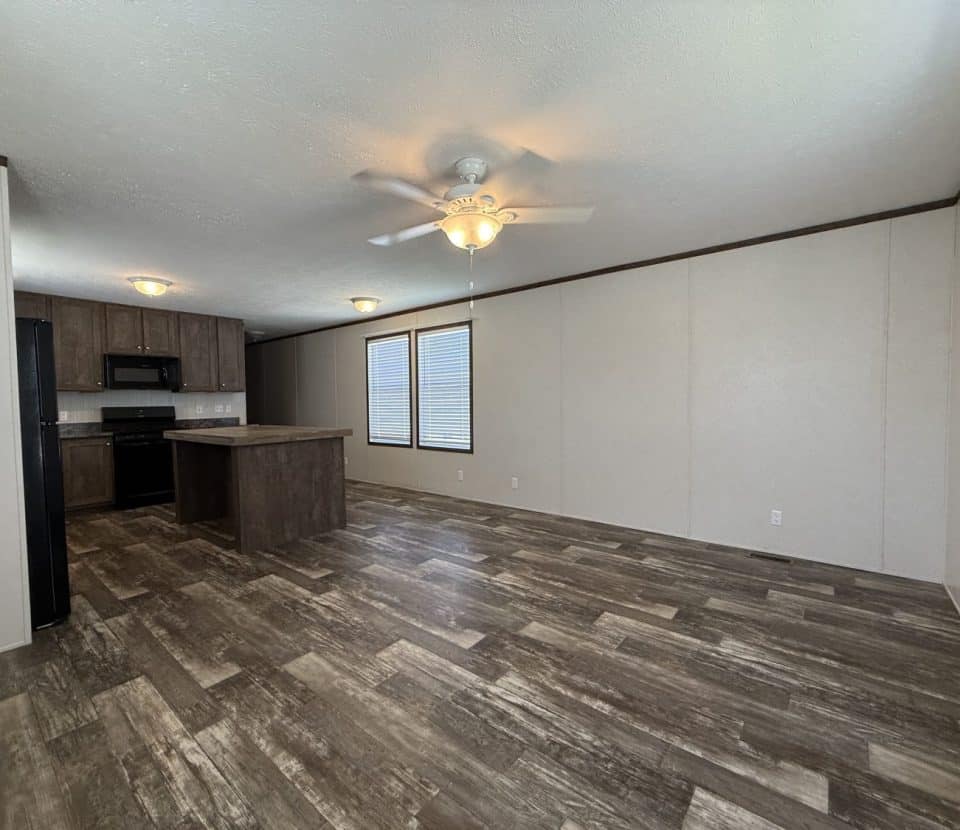 Open-concept kitchen and living area with wood-look flooring, beige walls, a ceiling fan with light, large windows, and dark wood cabinets and appliances in the kitchen.