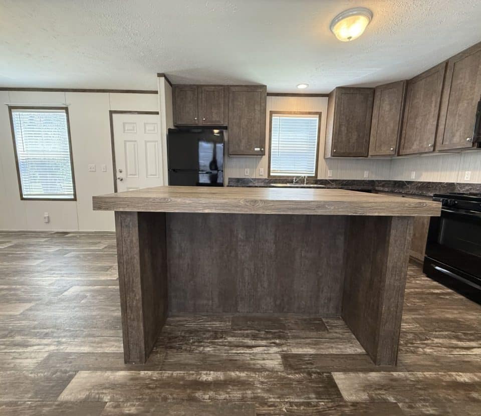A modern kitchen with dark wood cabinets, black appliances, a center island, and wood-style flooring. Windows and a white door are visible in the background, providing natural light.