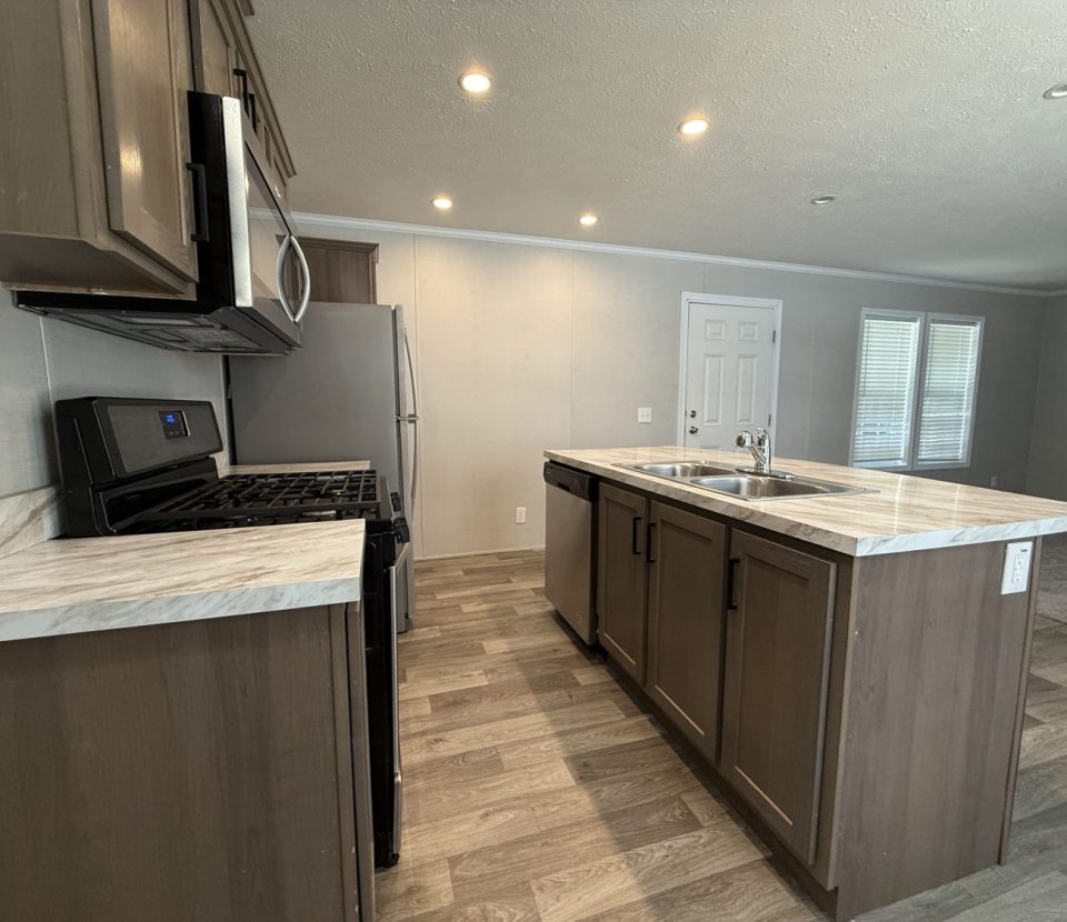 Modern kitchen with brown cabinets, marble countertops, stainless steel appliances, and a center island with a sink. The space is well-lit with recessed ceiling lights and has wood-patterned flooring.