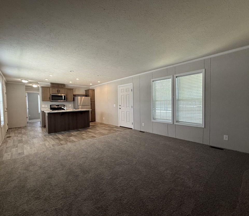 Empty modern living room with carpet flooring, large windows with blinds, and an open kitchen area featuring wood cabinets, stainless steel appliances, and vinyl flooring. White walls and ceiling with recessed lighting.