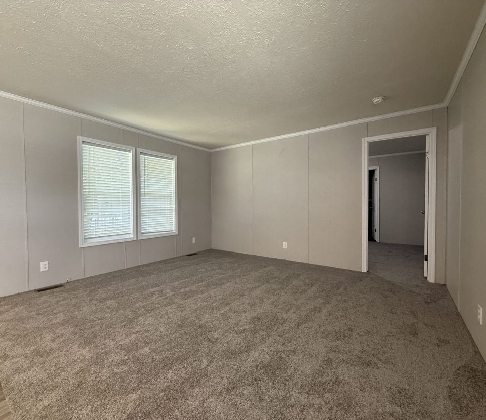 A spacious, empty room with beige carpet, light-colored walls, two windows with blinds, and two doorways leading to other rooms. The room is well-lit by natural light from the windows.