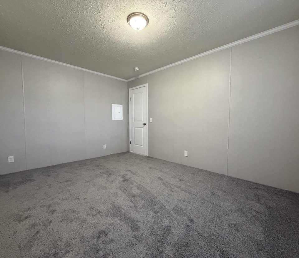 Empty room with light gray walls, white trim, a closed white door, a small access panel, a ceiling light fixture, and gray carpeted flooring. The room is well-lit and has a clean, minimal appearance.