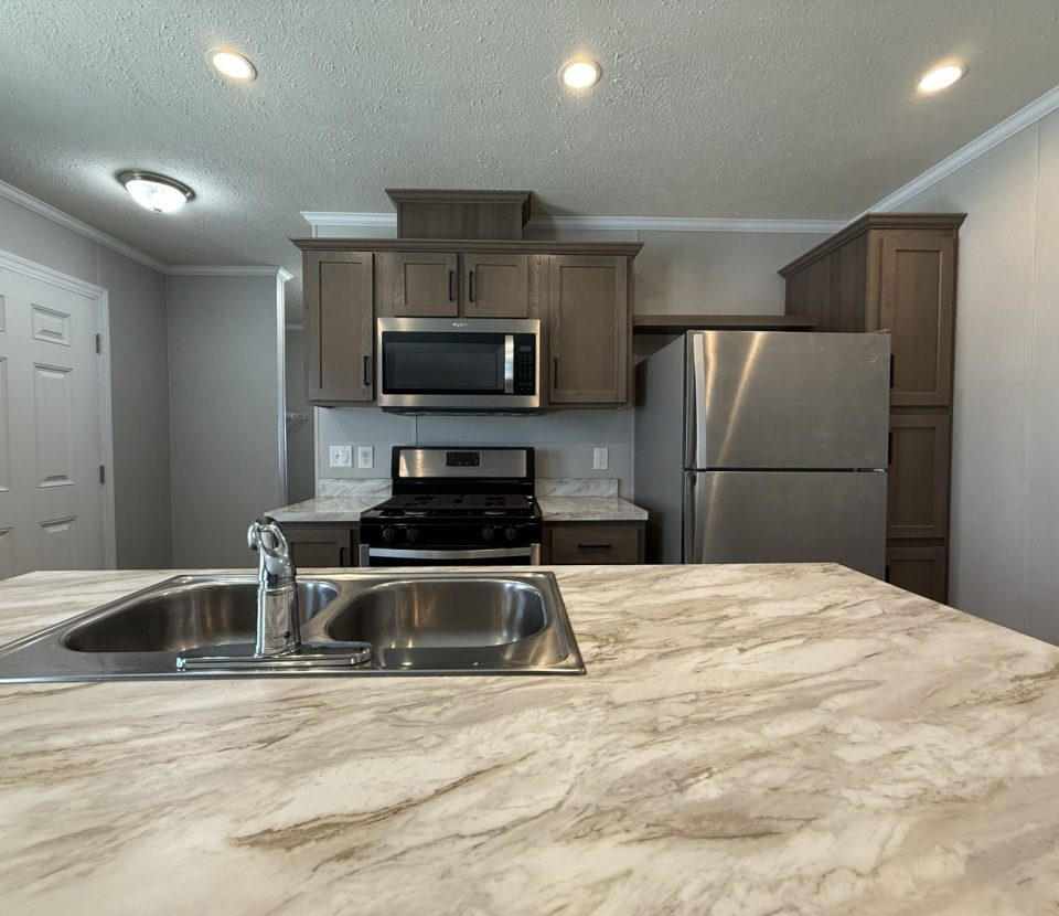 Modern kitchen with marble-look countertops, a double sink, stainless steel refrigerator, gas stove with oven, built-in microwave, brown cabinets, and recessed ceiling lights. White walls and door are visible in the background.