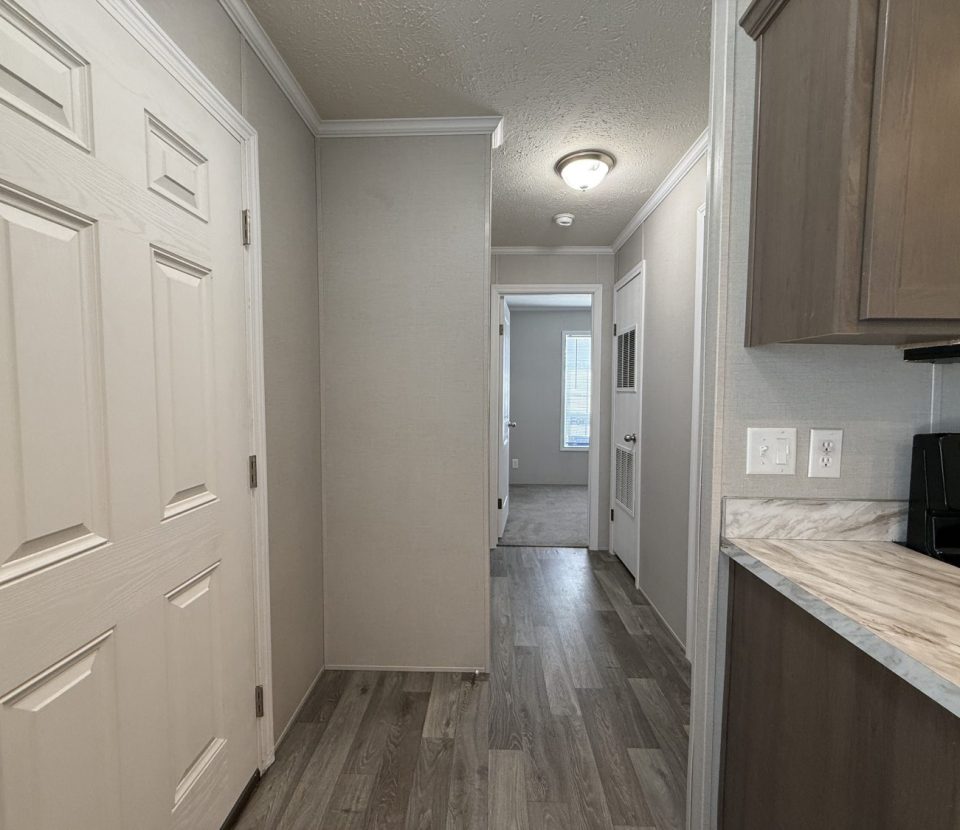 A hallway with wood-style flooring, a white front door on the left, light gray walls, and a kitchen area on the right with marble-patterned countertops and wooden cabinets. A light fixture is on the ceiling.