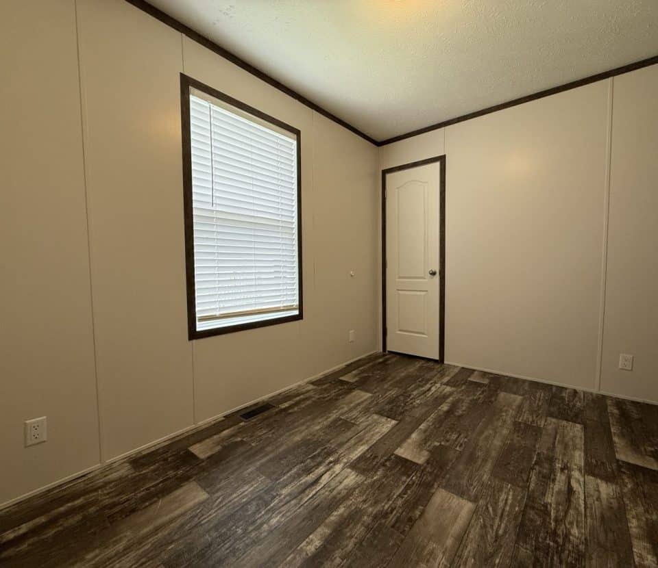 Empty room with beige walls, dark trim, wood-patterned vinyl flooring, a window with closed white blinds, and a white door with a round handle. The space is well-lit from overhead lighting.