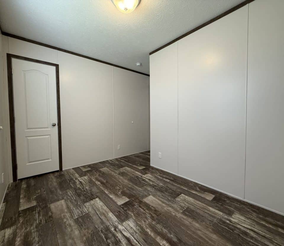Empty room with beige walls, a white ceiling light fixture, wood-look vinyl flooring, and a closed white door; sunlight comes in from a window partially visible on the left.