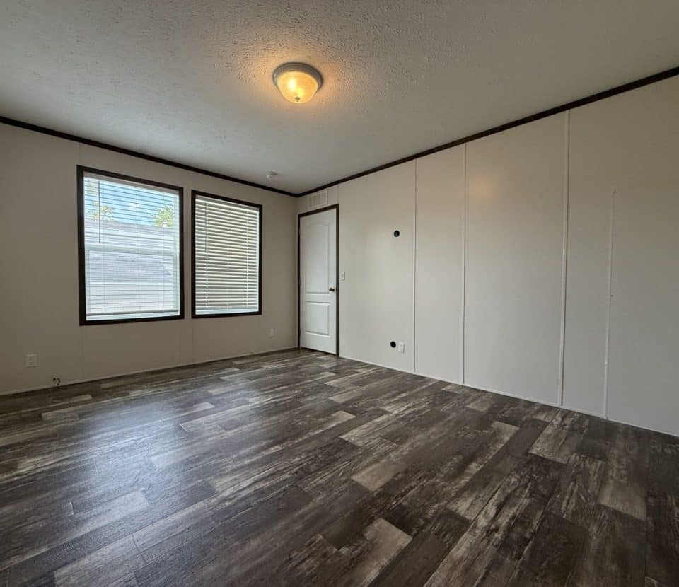An empty room with wood-look flooring, white walls, a white door, and three large windows letting in natural light. The ceiling has a central light fixture.