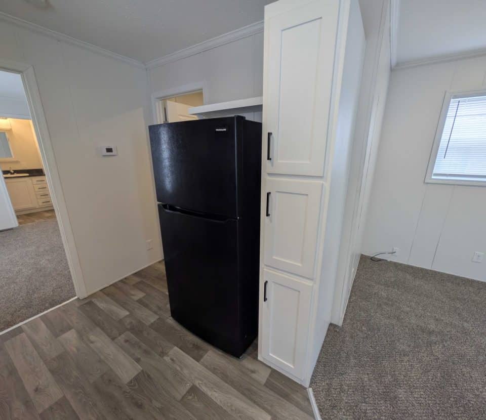 A black refrigerator stands next to white cabinets in a corner between two rooms with gray wood-style flooring and neutral walls; doorways and a window are visible in the background.