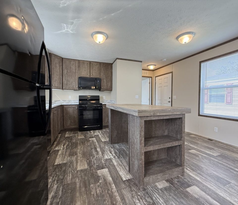 A modern kitchen with brown wood cabinets, a black refrigerator, stove, and microwave, a large island with open shelves, laminate flooring, and ample natural light from a window.