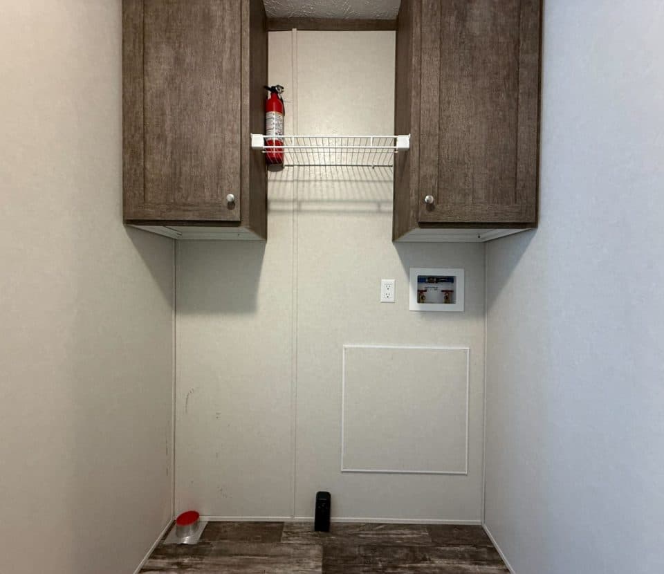 A small laundry room with two wooden cabinets, a white wire shelf holding a fire extinguisher, utility hookups on the wall, and a fire alarm panel. The floor is wood-patterned, with some items along the edges.