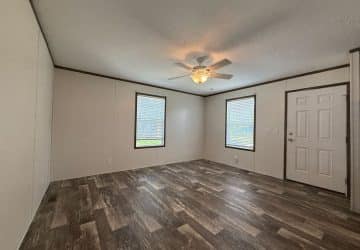 A spacious, empty room with wood-look flooring, beige walls, a white ceiling fan with a light, two large windows, and a white door. Natural light fills the room through the windows.
