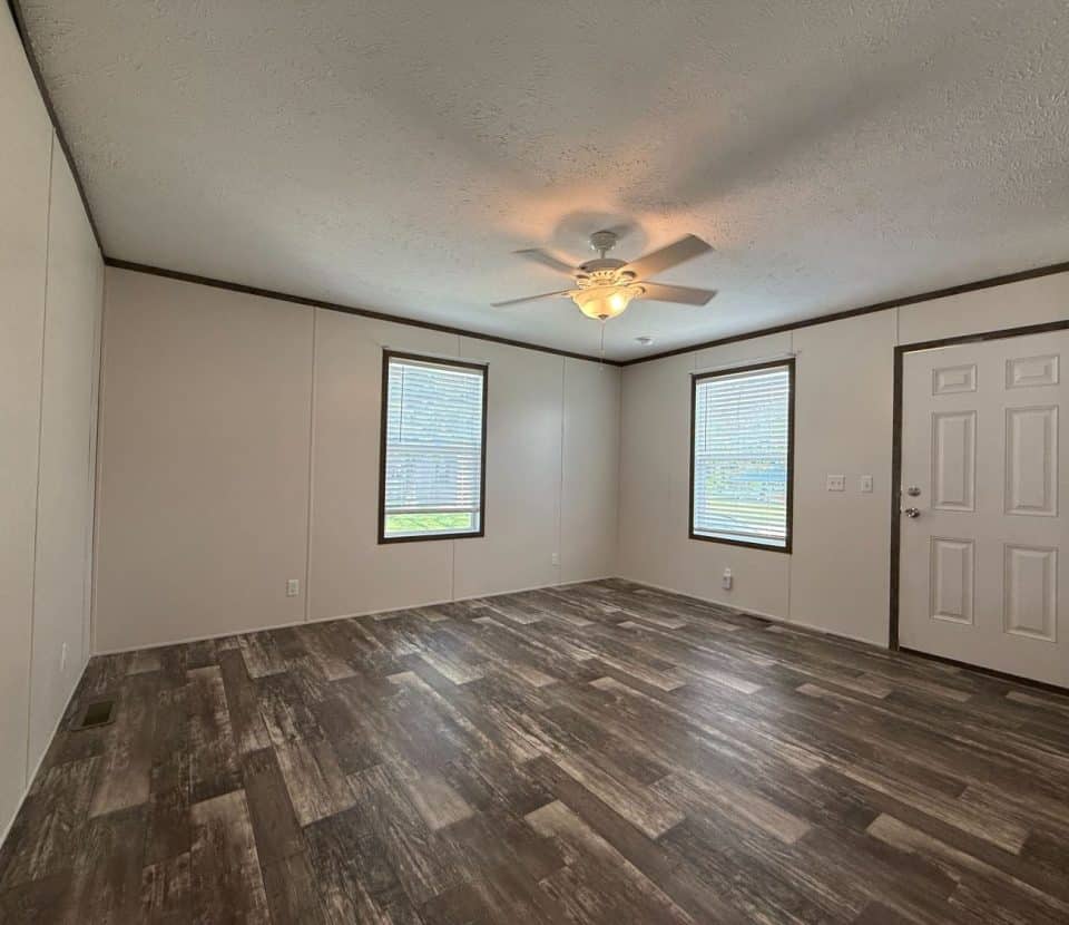 A spacious, empty room with wood-look flooring, beige walls, a white ceiling fan with a light, two large windows, and a white door. Natural light fills the room through the windows.
