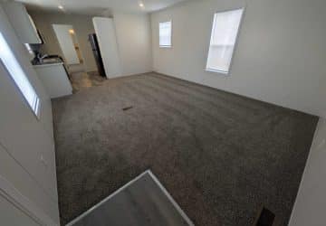 Empty room with gray carpet, white walls, and two windows letting in natural light. The space has an open layout with a kitchen area visible in the background and a small section of vinyl flooring in the foreground.