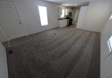 A spacious, unfurnished room with gray carpet, white walls, a front door, two tall windows, and an open kitchen area with white cabinets and stainless steel appliances in the back.