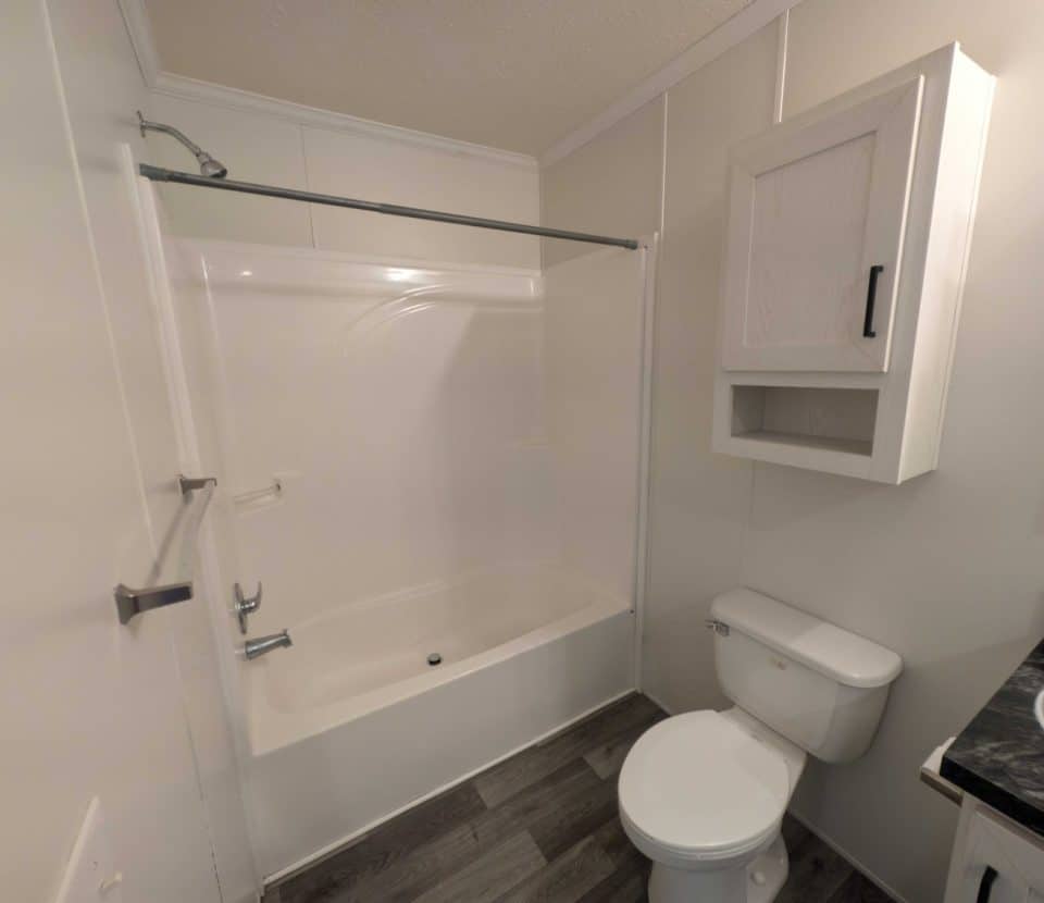 A small, modern bathroom with a white bathtub and shower, a toilet, a wall-mounted cabinet, and a dark countertop with a sink. The floor has gray wood-patterned vinyl planks.