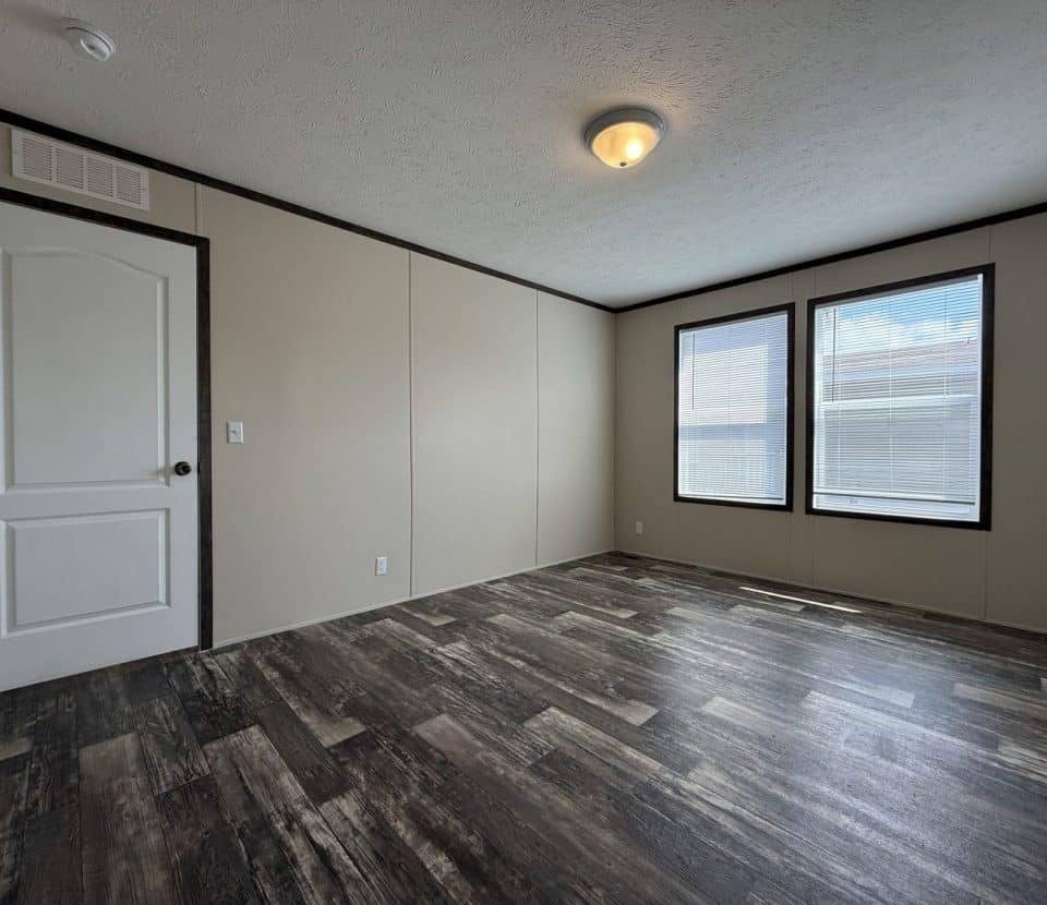 A bright, empty room with beige walls, dark wood-style flooring, a white door, and two large windows letting in natural light.