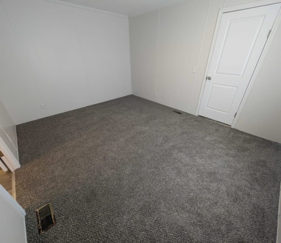 Empty room with light gray carpet, white walls, a white door, and a floor vent. The room is unfurnished and has a doorway leading to another area with light-colored flooring.