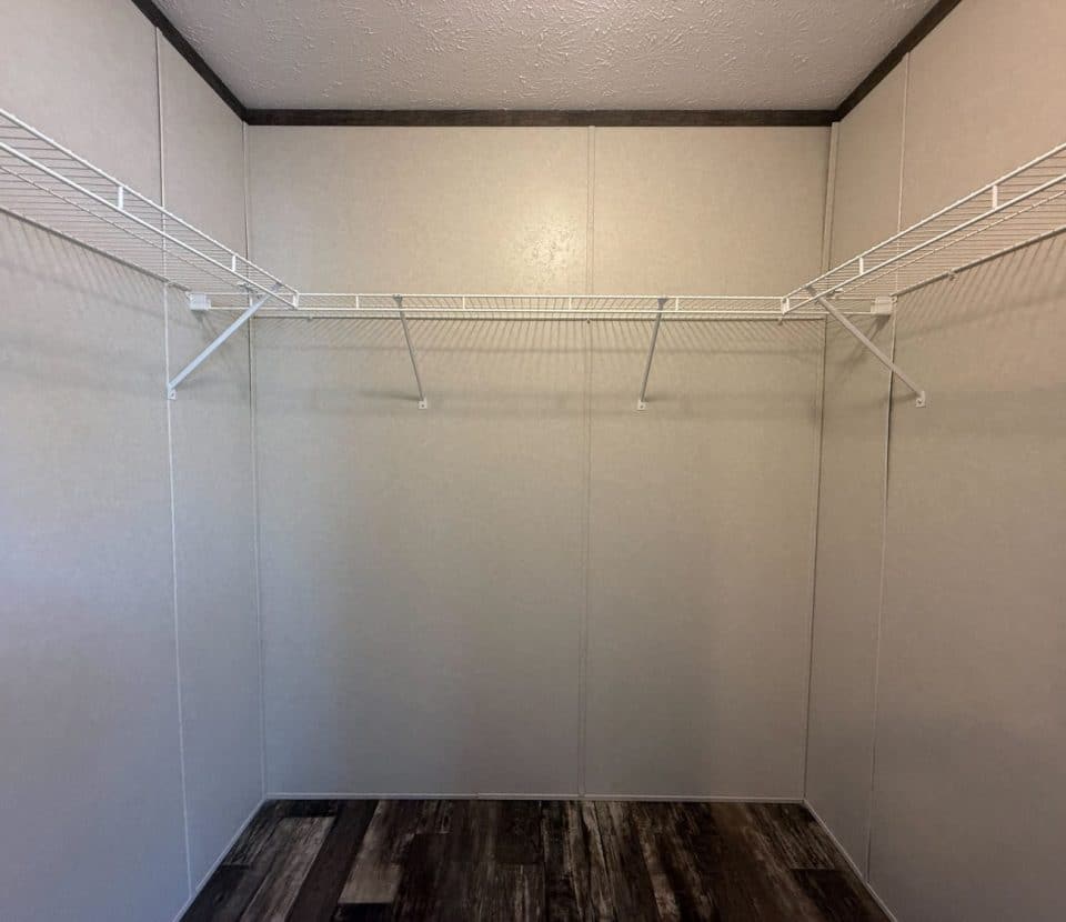 Empty walk-in closet with beige walls, dark wood floor, and two white wire shelves along the left and back walls. The closet is well-lit and uncluttered.