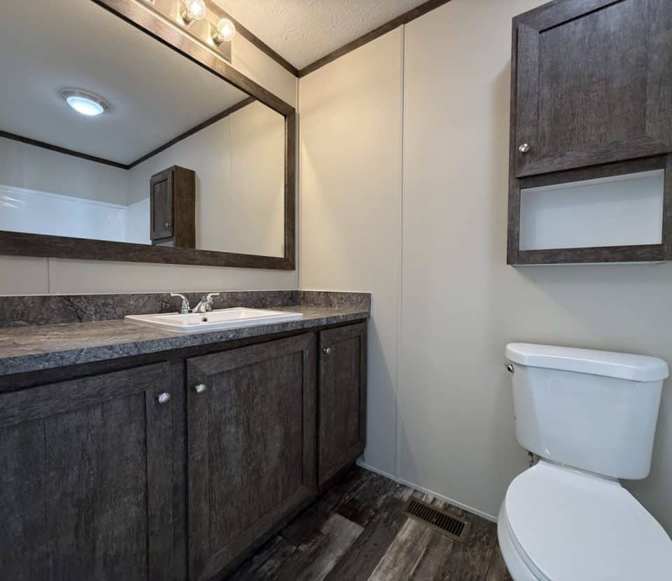 Modern bathroom with dark wood cabinets, a large mirror above a sink, a white toilet, a matching wall cabinet, and light-colored walls and flooring. The space is clean and uncluttered.