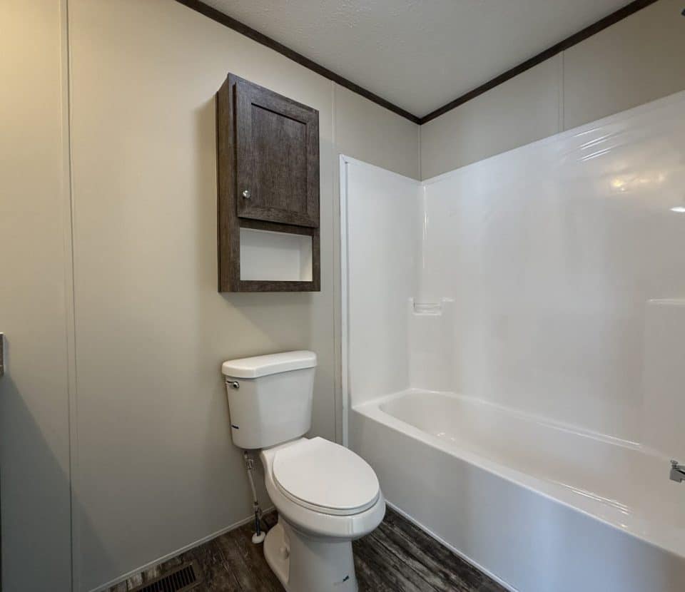 A clean, modern bathroom with a white toilet and bathtub-shower combo, wood-look flooring, beige walls, and a dark wooden cabinet mounted above the toilet.