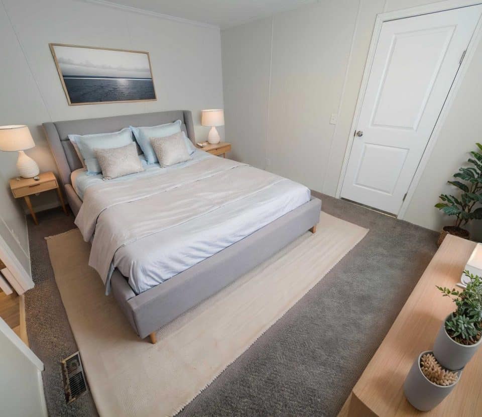 A modern bedroom with a gray upholstered bed, white bedding, two bedside tables with lamps, a potted plant, a dresser with succulents, and a serene landscape painting above the bed.