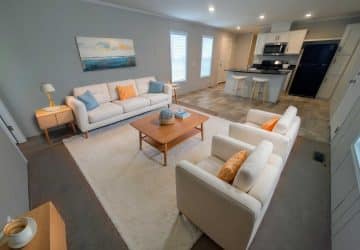 Modern living room and kitchen with beige sofas, two armchairs with orange pillows, a wooden coffee table, white rug, and light cabinets. The kitchen has black appliances and a breakfast bar with two stools.
