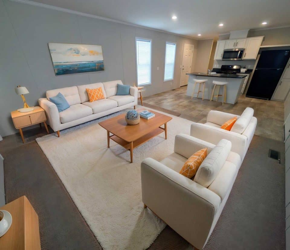 Modern living room and kitchen with beige sofas, two armchairs with orange pillows, a wooden coffee table, white rug, and light cabinets. The kitchen has black appliances and a breakfast bar with two stools.
