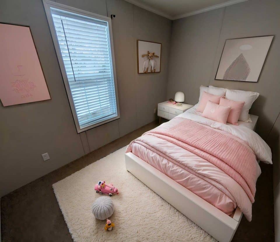 A neatly made bed with pink bedding and pillows sits on a white rug in a small, tidy bedroom. The walls are light gray, with framed artwork and a window with closed blinds. A nightstand holds a lamp and decor items.
