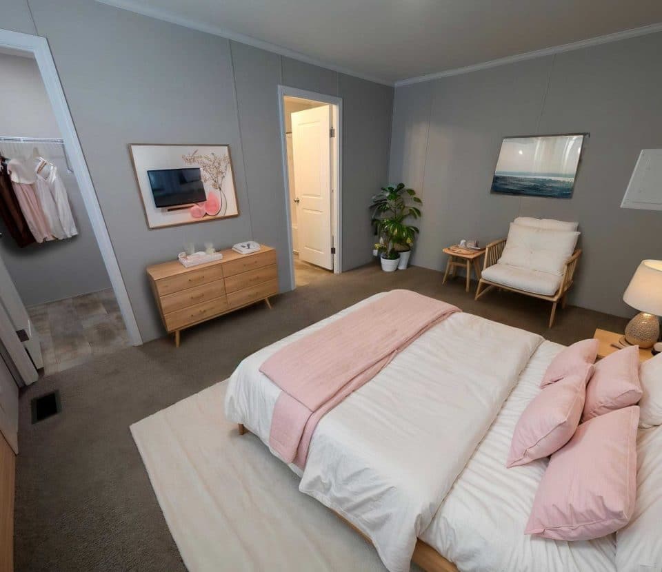 A modern bedroom features a neatly made bed with pink and white bedding, a wooden dresser and nightstand, a wall-mounted TV, a cozy chair, a potted plant, and an open walk-in closet with hanging clothes.