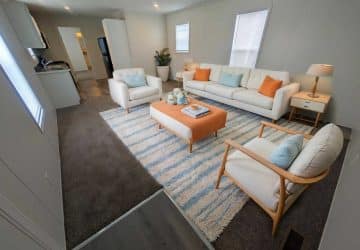 Modern living room with white walls, white sofa and armchairs, blue and orange accent pillows, orange ottoman, striped rug, side tables with lamps, plant, and kitchen visible in the background.