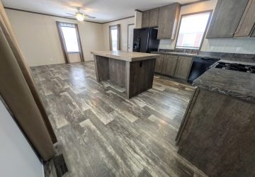 Spacious modern kitchen with wood-look flooring, dark wood cabinets, black appliances, a large center island, and two windows letting in natural light. Ceiling fan and curtains are visible in the background.